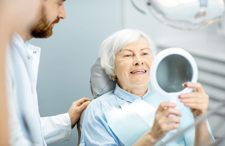 Happy elderly woman enjoying her beautiful toothy smile looking to the mirror in the dental office